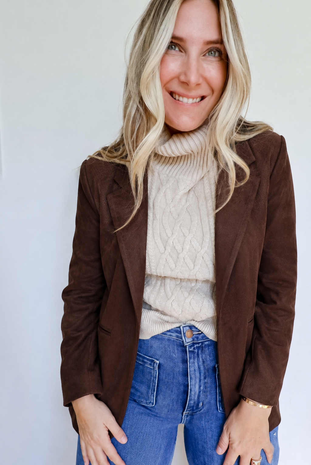 A woman wearing a dark brown suede blazer over a cream cable-knit turtleneck sweater and blue jeans, standing against a plain white wall.
