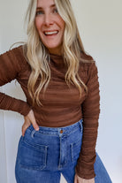 Woman wearing a brown shirred long sleeve mock neck top with blue jeans.