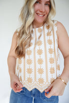 Woman wearing a white sleeveless top with gold patterns and blue jeans against a white background